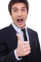 Thumb up for success Happy young man in formalwear showing his thumb up and keeping mouth open while standing isolated on white background photo