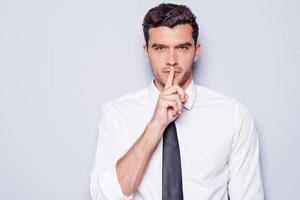 Keep silence Handsome young man in white shirt looking at camera and holding finger on lips while standing against grey background photo