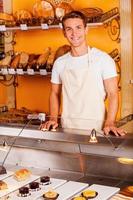 How may I help you Handsome young man in apron looking at camera and smiling while standing in bakery shop photo