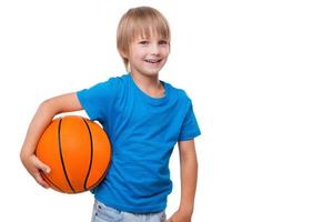 Me encanta el baloncesto, un niño alegre que sostiene una pelota de baloncesto y sonríe mientras está de pie, aislado en blanco. foto