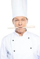 What to cook Surprised mature chef in white uniform holding wooden spoon in his mouth while standing against white background photo