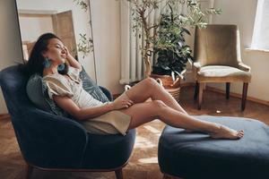 Beautiful young woman keeping eyes closed while relaxing in comfortable chair at home photo