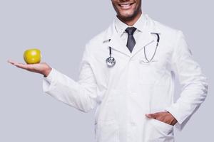 Eat healthy Cropped image of African doctor stretching out green apple while standing against grey background photo