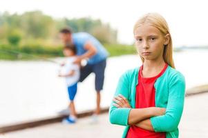 la pesca es demasiado aburrida para mí, una niña deprimida que mantiene los brazos cruzados y mira la cámara mientras un hombre con un niño pequeño pesca en el fondo foto