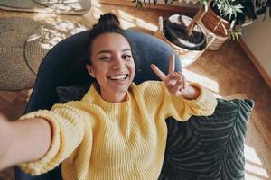 Top view of happy young woman making selfie and gesturing while relaxing in chair at home photo