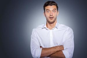 Wow Surprised young man in white shirt staring at camera and keeping arms crossed while standing against grey background photo