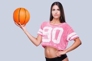 Are you ready to play Beautiful young female cheerleader holding basketball ball and looking at camera while standing against grey background photo