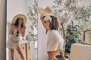 mujer joven juguetona con sombrero elegante y sonriendo mientras está de pie frente al espejo en casa foto