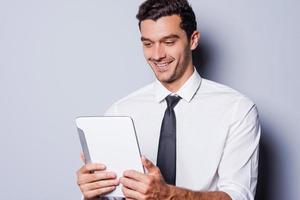 Good news Cheerful young man in shirt and tie looking at digital tablet and smiling while standing against grey background photo