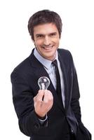 Got idea Cheerful young man in formalwear stretching out hand with light bulb and smiling while standing isolated on white background photo