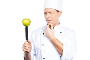 cocinar comida vegetariana. chef maduro confiado en uniforme blanco sosteniendo un cuchillo de cocina con manzana verde en la parte superior y mirándolo mientras está de pie contra el fondo blanco foto