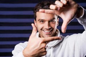 Focus on me Playful young man in white sweater gesturing finger frame and smiling while standing against striped background photo