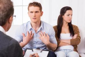 It is not my fault Displeased young man talking to psychiatrist and gesturing while his wife sitting near him and keeping arms crossed photo