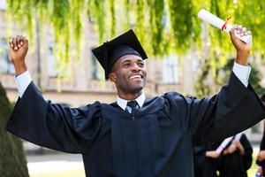 I have finally graduated Happy young African man in graduation gowns holding diploma and rising arms up while his friends standing in the background photo