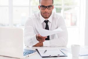 I have some paperwork for you Confident young African man in shirt and tie stretching out documents and looking at you while sitting at his working place photo