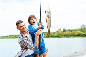 It is so big Father and son stretching a fishing rod with fish on the hook while little boy looking excited and keeping mouth open photo