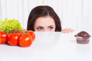 What to choose Thoughtful young woman looking out of the table while fresh vegetables and chocolate muffin laying on it photo