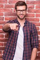 Te elijo a ti, joven apuesto, apuntándote y sonriendo mientras estás de pie contra la pared de ladrillos. foto