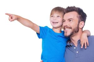 ¿Qué hay allí? Vista lateral de feliz padre e hijo mirando hacia otro lado mientras el niño señala hacia otro lado y ambos parados aislados en blanco. foto