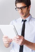 It is just amazing Surprised young man in shirt and tie looking at digital tablet and keeping mouth open while standing against grey background photo