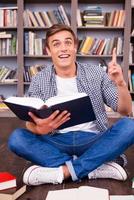 Bingo Happy young man holding book and gesturing while sitting against bookshelf