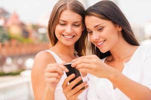 Look at this Two beautiful young women looking at mobile phone and smiling while standing outdoors together photo