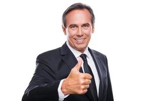 This is success Cheerful mature man in formalwear looking at camera and smiling while showing his thumb up and standing against white background photo