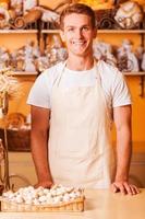 How may I help you Handsome young man in apron looking at camera and smiling while standing in bakery shop photo