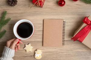 Woman hand holding black coffee cup with blank notebook and Christmas cookies on table. Xmas, Happy New Year, Goals, Resolution, To do list, Strategy and Plan concept photo