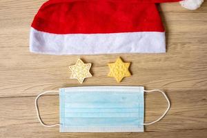 Merry Christmas with homemade cookies, Santa clause hat and virus protective face mask on wood table background. Health, Covid 19 pandemic, Xmas, party, holiday and happy New Year concept photo