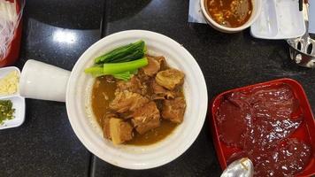 vista superior de costillas de cerdo estofadas con col rizada en plato caliente e hígado fresco en bandeja roja y guarnición con ají verde en rodajas y ajo en mesa de comedor negra. fideos de arroz con salsa picante o agria. foto