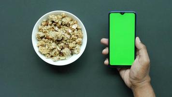 vista aérea del tazón de granola junto a la mano de una persona que sostiene un teléfono inteligente con pantalla verde video