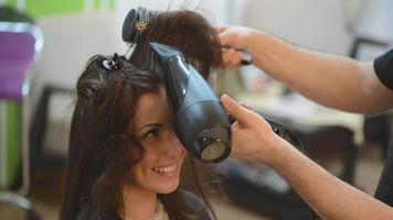 Man hairdresser drying long brown hair of a customer woman with round brush. video