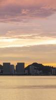 vertikal Timelapse silhuett se av vid vatten stad med höghus skyskrapa byggnad nära hav kust strand medan solnedgång solnedgång tid med vanilj skymning himmel video