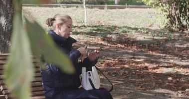 belle fille dans le parc sur un banc avec un sac blanc regarde le téléphone. video