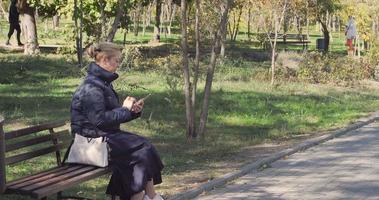 linda garota no parque em um banco com uma bolsa branca Olha para o telefone video