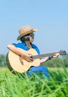 las mujeres de pelo corto usan sombrero y gafas de sol sentadas tocando la guitarra en el campo de hierba foto