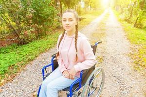 Young happy handicap woman in wheelchair on road in hospital park waiting for patient services. Paralyzed girl in invalid chair for disabled people outdoor in nature. Rehabilitation concept. photo