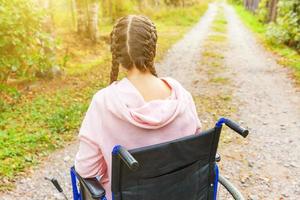 joven discapacitada feliz en silla de ruedas en la carretera en el parque del hospital esperando los servicios del paciente. niña paralizada en silla inválida para personas discapacitadas al aire libre en la naturaleza. concepto de rehabilitación. foto