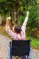 joven discapacitada feliz en silla de ruedas en la carretera en el parque del hospital disfrutando de la libertad. niña paralizada en silla inválida para personas discapacitadas al aire libre en la naturaleza. concepto de rehabilitación. foto