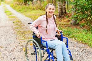 Young happy handicap woman in wheelchair on road in hospital park enjoying freedom. Paralyzed girl in invalid chair for disabled people outdoor in nature. Rehabilitation concept. photo