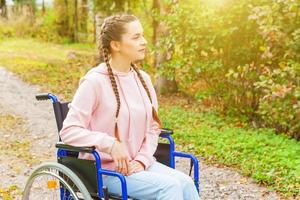 Young happy handicap woman in wheelchair on road in hospital park waiting for patient services. Paralyzed girl in invalid chair for disabled people outdoor in nature. Rehabilitation concept. photo