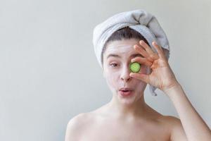 Beauty portrait of woman in towel on head with white nourishing mask or creme on face holding cucumber slices, white background isolated photo