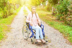 Young happy handicap woman in wheelchair on road in hospital park waiting for patient services. Paralyzed girl in invalid chair for disabled people outdoor in nature. Rehabilitation concept. photo