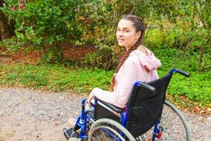 Young happy handicap woman in wheelchair on road in hospital park waiting for patient services. Paralyzed girl in invalid chair for disabled people outdoor in nature. Rehabilitation concept. photo