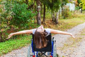 joven discapacitada feliz en silla de ruedas en la carretera en el parque del hospital disfrutando de la libertad. niña paralizada en silla inválida para personas discapacitadas al aire libre en la naturaleza. concepto de rehabilitación. foto