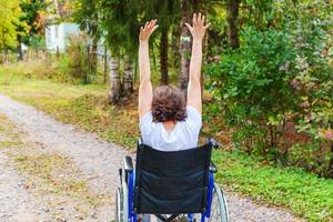 Young happy handicap woman in wheelchair on road in hospital park enjoying freedom. Paralyzed girl in invalid chair for disabled people outdoor in nature. Rehabilitation concept. photo