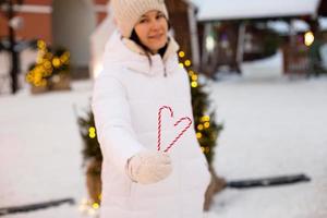mujer sostiene en sus manos un bastón de caramelo en forma de corazón al aire libre con ropa blanca de abrigo en el mercado festivo de invierno. Guirnaldas de luces de hadas decoradas ciudad de nieve para año nuevo. humor navideño foto