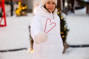 Woman holds in his hands candy cane in the shape of a heart outdoor in white warm clothes in winter festive market. Fairy lights garlands decorated snow town for new year. Christmas mood photo