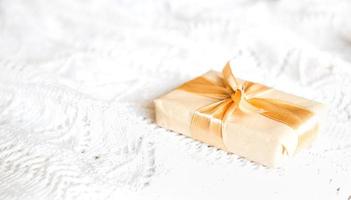 Box with Christmas gift in light paper is tied with a gold ribbon on white knitted background with decor. Golden garland of stars. New Year. Congratulations, surprise. space for text photo